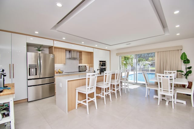 kitchen with a raised ceiling, wall chimney exhaust hood, stainless steel fridge with ice dispenser, and white cabinets