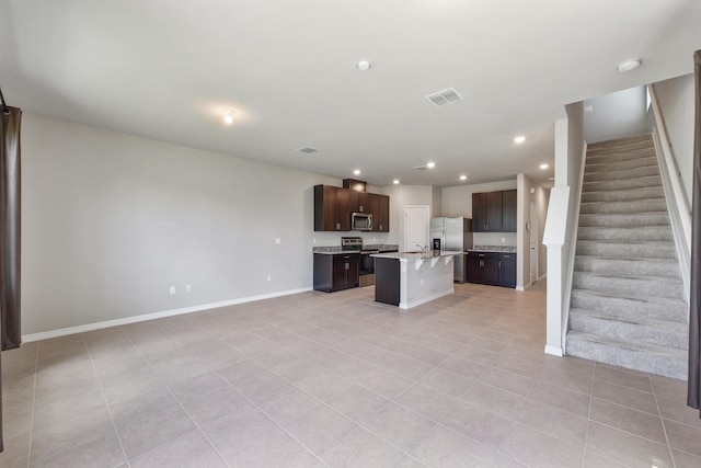 kitchen with light tile patterned flooring, a center island with sink, appliances with stainless steel finishes, a kitchen bar, and dark brown cabinets