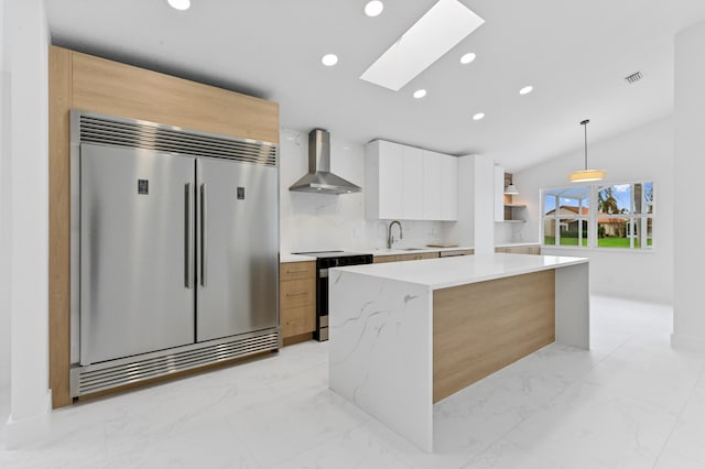 kitchen featuring built in refrigerator, white cabinets, range, wall chimney range hood, and vaulted ceiling with skylight