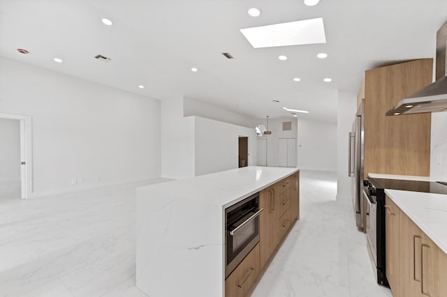 kitchen with a skylight, wall chimney range hood, light stone countertops, a spacious island, and appliances with stainless steel finishes