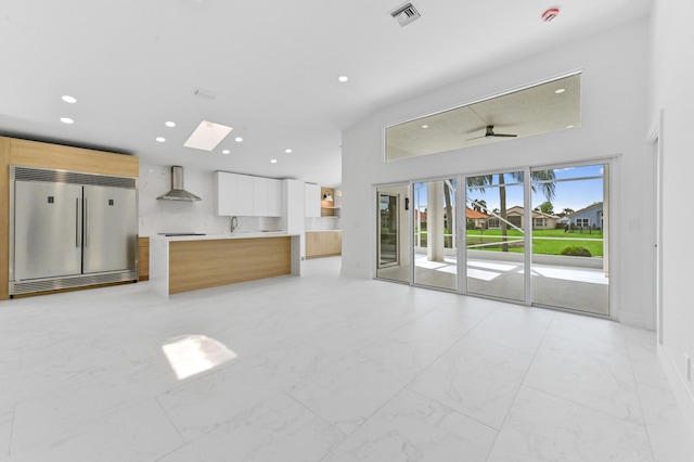 kitchen with white cabinets, stainless steel built in refrigerator, a skylight, wall chimney exhaust hood, and ceiling fan