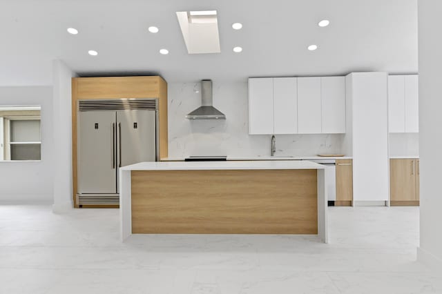 kitchen with tasteful backsplash, built in fridge, white cabinetry, wall chimney range hood, and a skylight