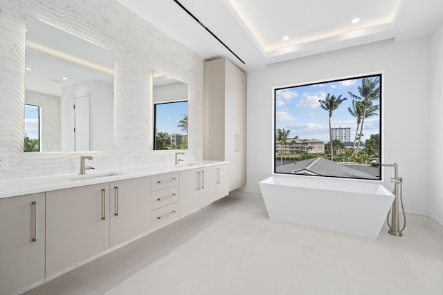 full bath with double vanity, decorative backsplash, a tray ceiling, a freestanding bath, and a sink