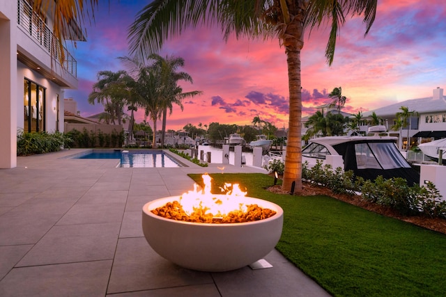 pool at dusk featuring a fire pit, an outdoor pool, a boat dock, a yard, and a patio area