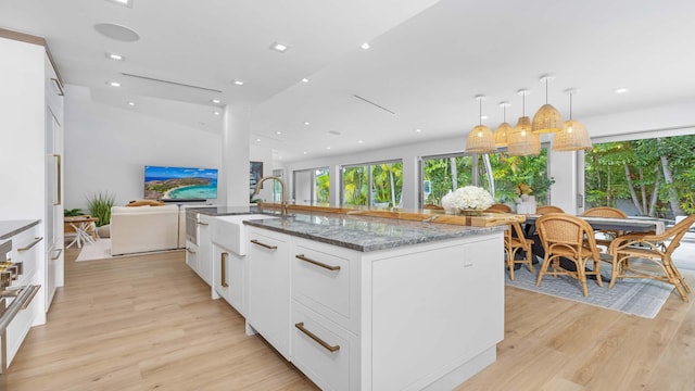 kitchen with hanging light fixtures, dark stone counters, light hardwood / wood-style floors, a kitchen island with sink, and white cabinets