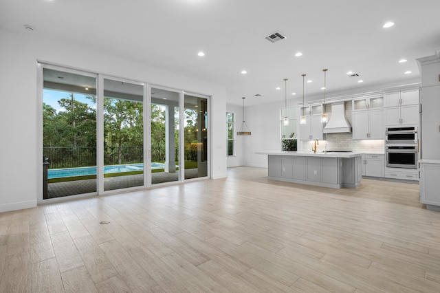 kitchen with custom exhaust hood, stainless steel double oven, a center island with sink, light hardwood / wood-style flooring, and hanging light fixtures