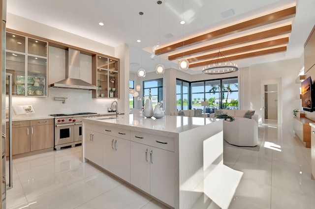 kitchen with white cabinetry, wall chimney exhaust hood, pendant lighting, range with two ovens, and a kitchen island with sink