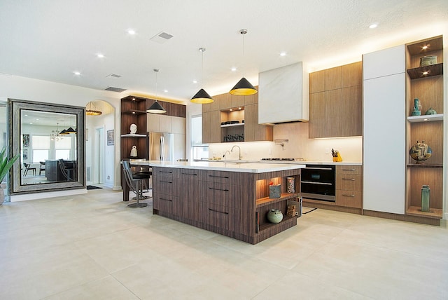 kitchen with sink, custom range hood, stainless steel refrigerator, pendant lighting, and a large island with sink