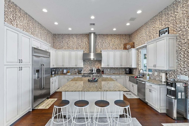 kitchen featuring wall chimney exhaust hood, a kitchen island, light stone countertops, white cabinetry, and appliances with stainless steel finishes