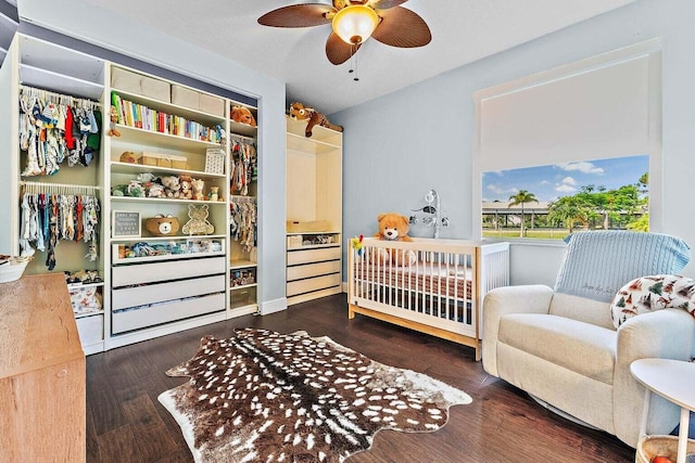 bedroom with a nursery area, dark wood-type flooring, ceiling fan, and a closet