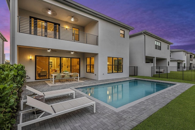 back house at dusk featuring ceiling fan, a balcony, a fenced in pool, and a patio