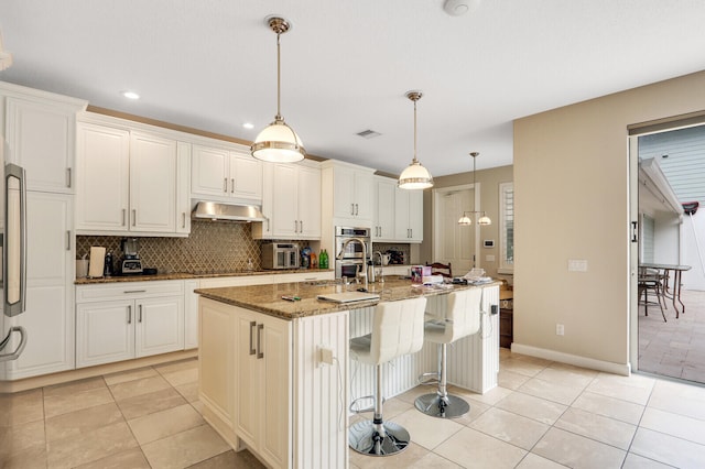 kitchen with stainless steel double oven, range hood, decorative light fixtures, stone countertops, and an island with sink