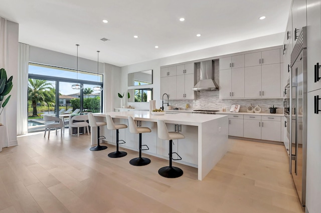 kitchen featuring backsplash, wall chimney range hood, hanging light fixtures, light wood-type flooring, and an island with sink