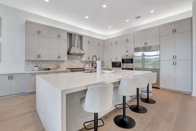 kitchen with wall chimney exhaust hood, a spacious island, built in appliances, light hardwood / wood-style flooring, and a breakfast bar area