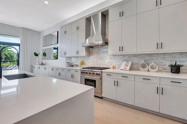 kitchen featuring luxury stove, wall chimney range hood, sink, decorative backsplash, and light wood-type flooring