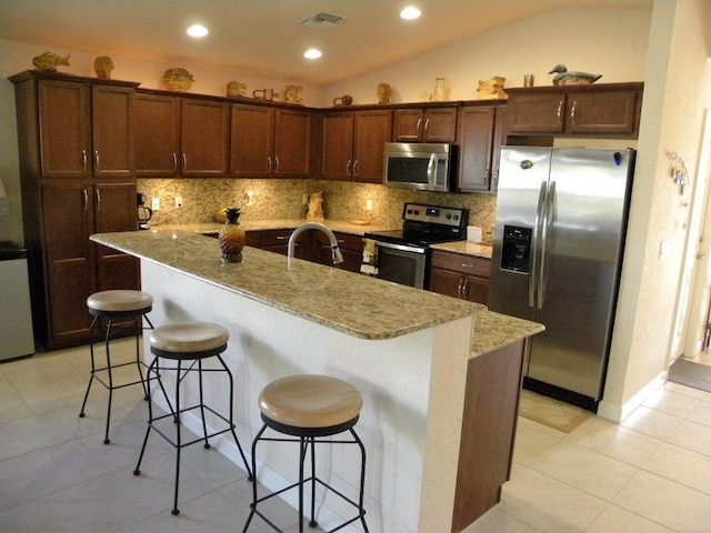 kitchen featuring stainless steel appliances, a kitchen bar, light stone countertops, vaulted ceiling, and an island with sink