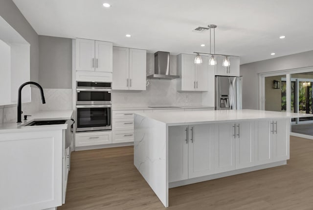 kitchen with pendant lighting, wall chimney exhaust hood, a kitchen island, white cabinetry, and appliances with stainless steel finishes