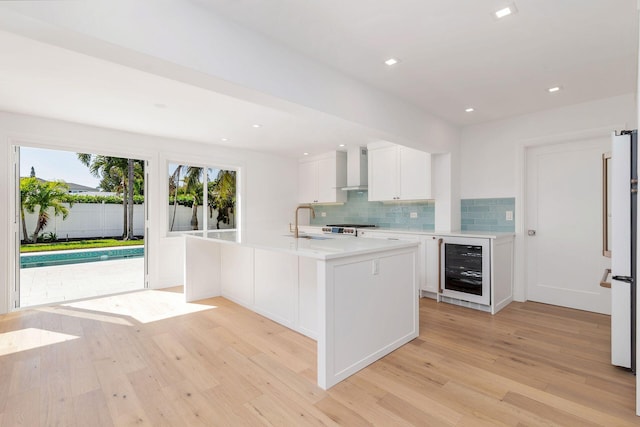 kitchen featuring wall chimney range hood, white refrigerator, an island with sink, white cabinets, and beverage cooler