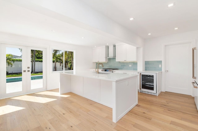 kitchen with white cabinetry, wine cooler, a kitchen island with sink, wall chimney range hood, and light hardwood / wood-style flooring