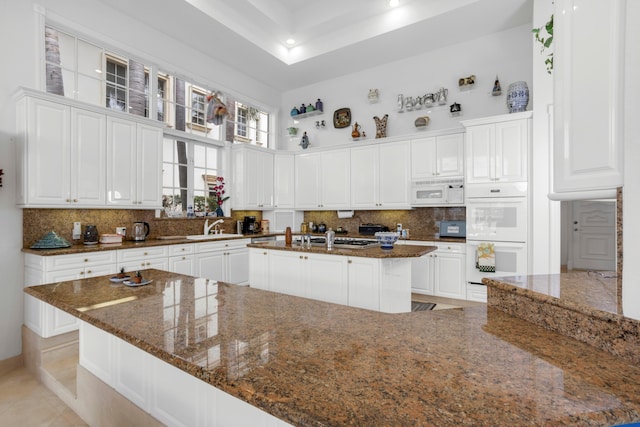 kitchen featuring a center island with sink, white cabinets, dark stone counters, and white appliances