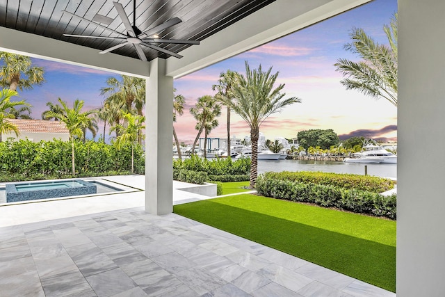 patio terrace at dusk with a swimming pool with hot tub, a water view, ceiling fan, and a lawn