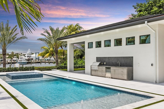 pool at dusk with a patio area, area for grilling, a water view, and exterior kitchen