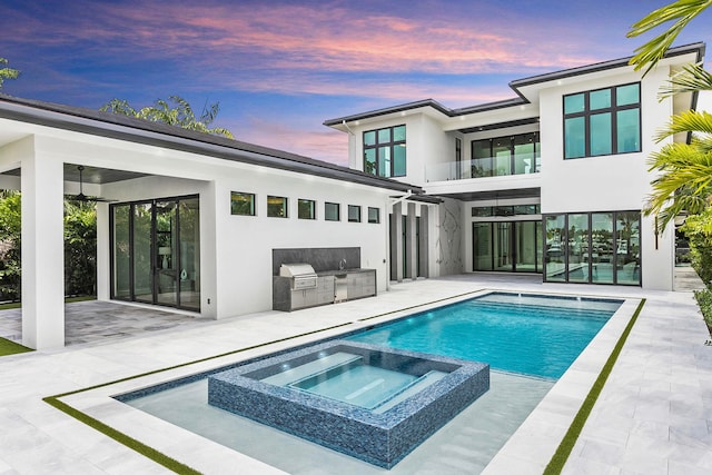 back house at dusk featuring an outdoor kitchen, a pool with hot tub, a patio, and a balcony