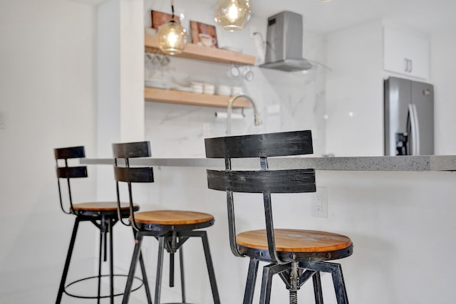 kitchen featuring white cabinets, a kitchen breakfast bar, stainless steel refrigerator, and wall chimney range hood