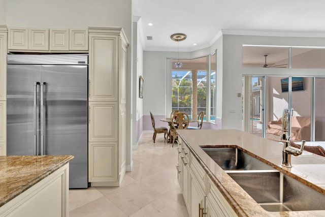 kitchen featuring stainless steel built in fridge, sink, cream cabinetry, decorative light fixtures, and light stone counters