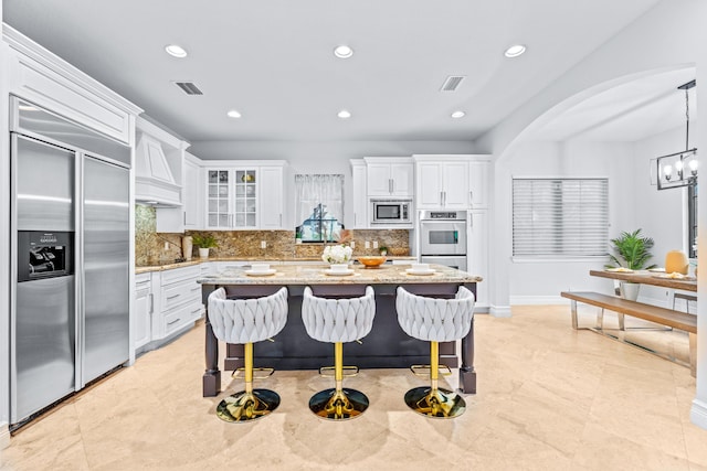 kitchen with light stone counters, backsplash, built in appliances, hanging light fixtures, and white cabinets