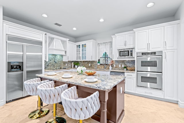 kitchen with a kitchen island, light stone counters, built in appliances, and white cabinets