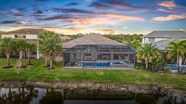 back house at dusk featuring a lawn, glass enclosure, a pool with hot tub, a water view, and a patio