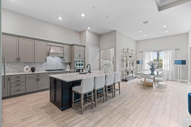 kitchen with a breakfast bar area, an island with sink, gray cabinets, custom range hood, and double oven