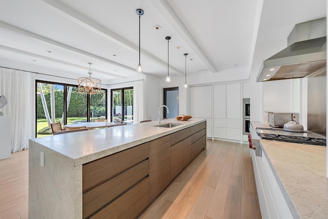 kitchen featuring a large island with sink, sink, wall chimney exhaust hood, light wood-type flooring, and decorative light fixtures