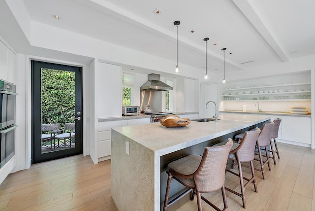 kitchen featuring white cabinetry, wall chimney exhaust hood, a kitchen breakfast bar, an island with sink, and decorative light fixtures