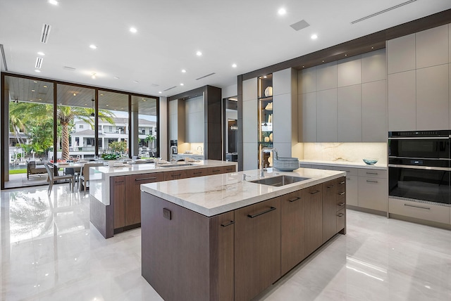 kitchen with light stone countertops, backsplash, stainless steel double oven, sink, and an island with sink