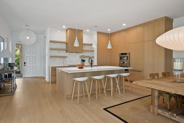 kitchen with sink, hanging light fixtures, decorative backsplash, a center island with sink, and light wood-type flooring