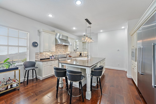 kitchen featuring an island with sink, built in appliances, sink, wall chimney range hood, and pendant lighting