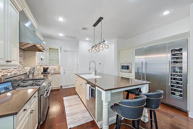 kitchen with built in appliances, sink, wall chimney range hood, dark hardwood / wood-style flooring, and a kitchen island with sink