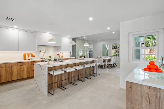 kitchen featuring white cabinetry, a kitchen bar, hanging light fixtures, a kitchen island with sink, and light stone counters