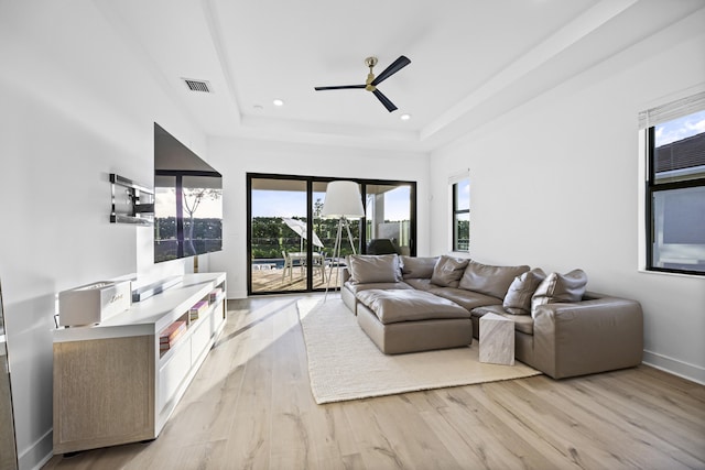 living room featuring light wood-type flooring, a raised ceiling, ceiling fan, and a healthy amount of sunlight