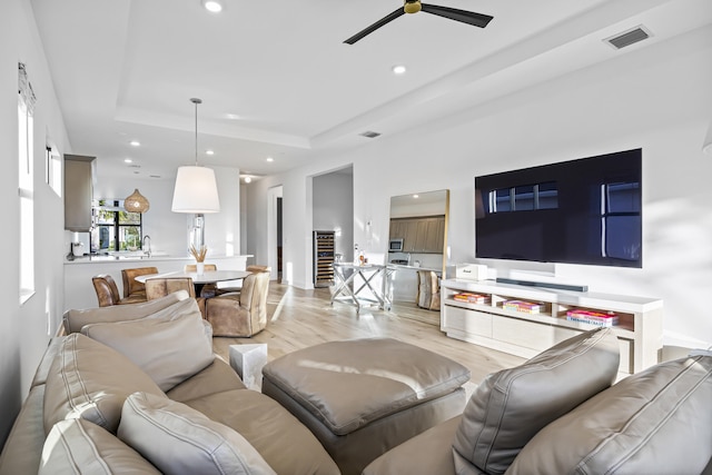 living room with ceiling fan, a tray ceiling, light wood-type flooring, and sink