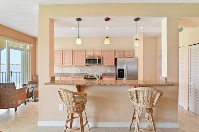 kitchen with light stone countertops, hanging light fixtures, stainless steel appliances, kitchen peninsula, and light brown cabinetry