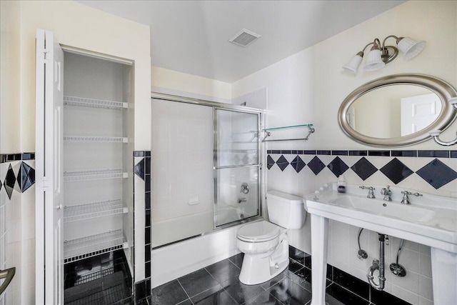 bathroom featuring tile patterned floors, bath / shower combo with glass door, tile walls, and toilet