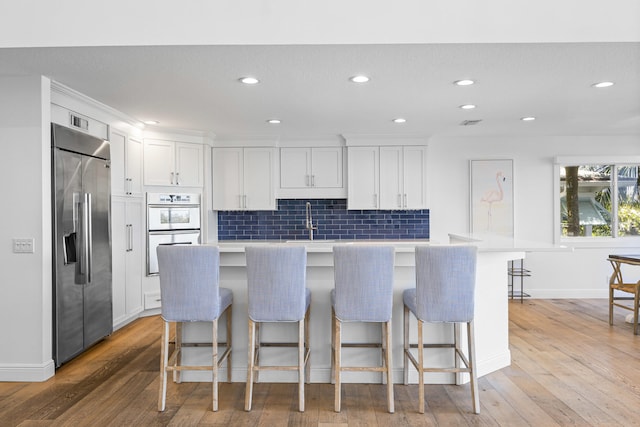 kitchen with white cabinetry, built in fridge, double oven, wood-type flooring, and a kitchen bar