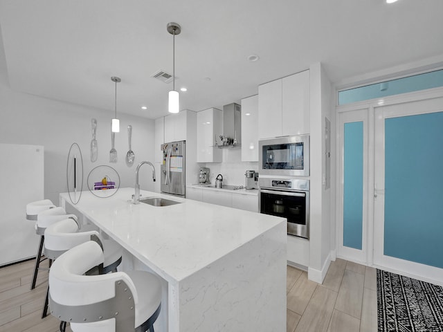 kitchen featuring stainless steel appliances, wall chimney range hood, sink, decorative light fixtures, and white cabinets