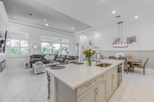 kitchen with ornamental molding, ceiling fan with notable chandelier, sink, hanging light fixtures, and an island with sink