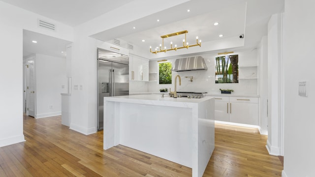 kitchen with an island with sink, built in refrigerator, white cabinets, wall chimney range hood, and light wood-type flooring