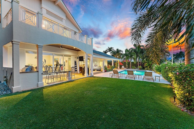 back house at dusk featuring an outdoor kitchen, a balcony, a patio area, and a lawn