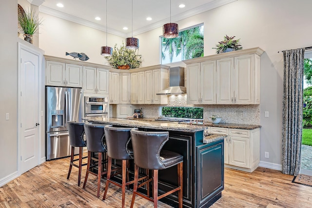 kitchen with wall chimney exhaust hood, stainless steel appliances, dark stone countertops, light hardwood / wood-style floors, and a kitchen island